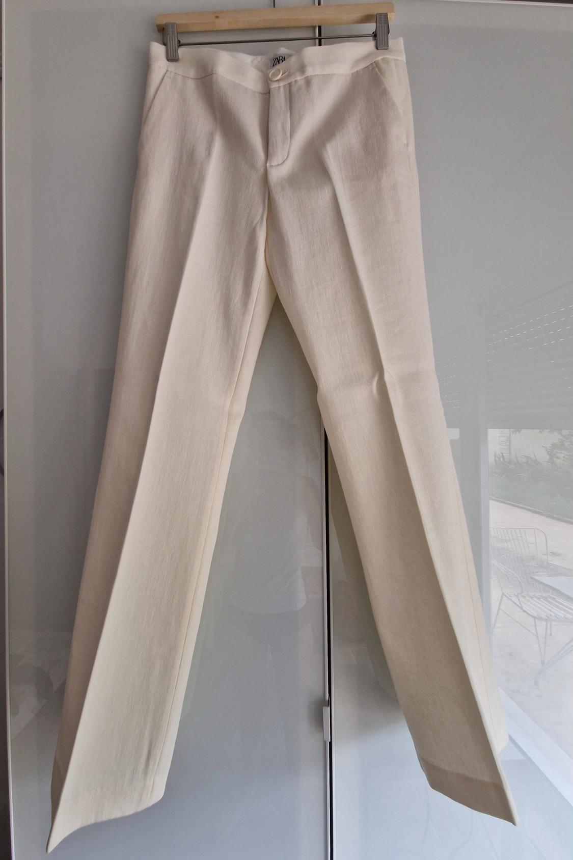 Light smart trousers with a front crease, hanging on a hanger over a glossy wardrobe. Inner label with the text Zara.