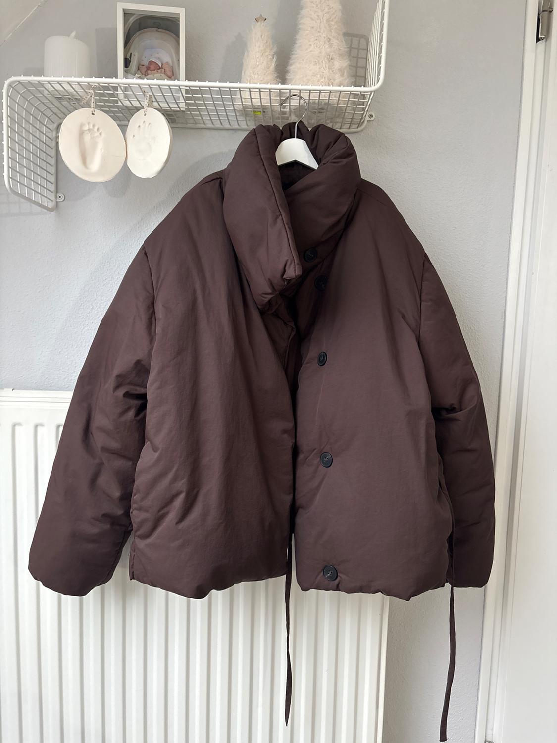 Brown padded jacket hanging on a white hanger. Background with a wire shelf, decorations and a framed photo.