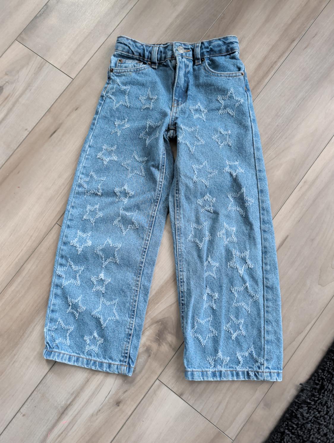 Light blue denim trousers with a white star print, laid on a light wooden floor.