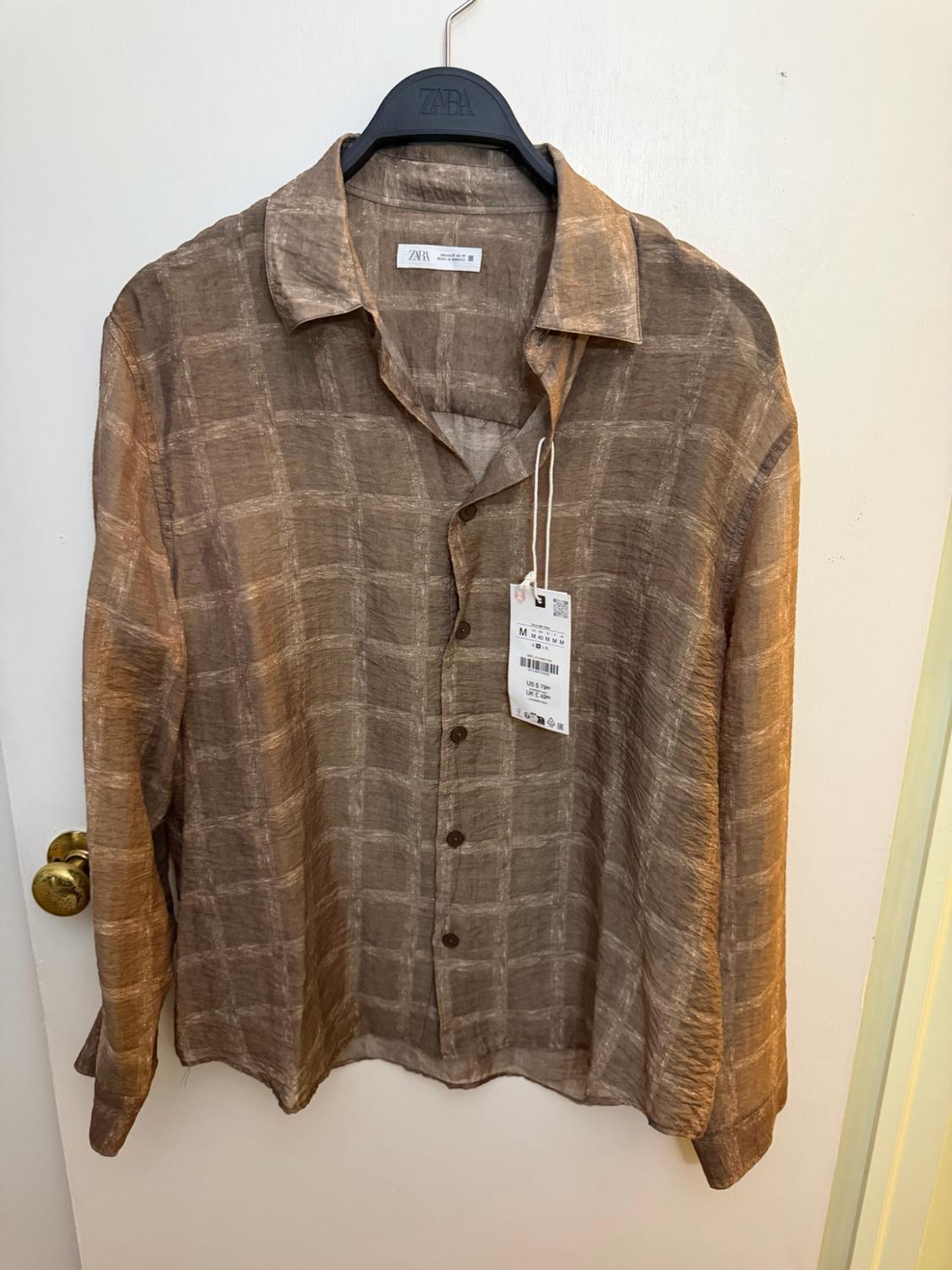 Brown long-sleeve plaid shirt hanging on a black hanger on a white door with a gold knob. ZARA text visible.