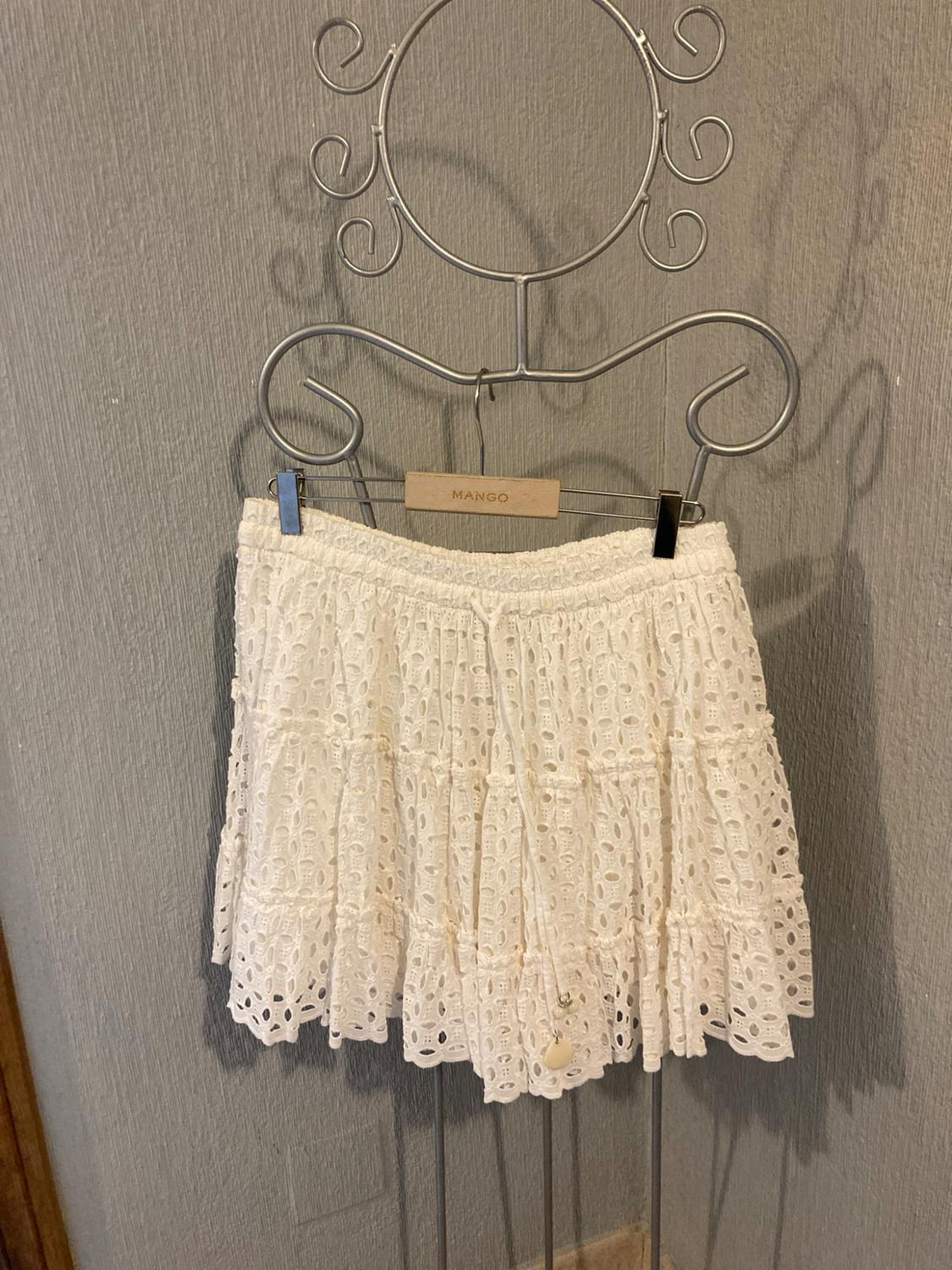 Short white skirt with eyelet embroidery and drawstring, hanging on a metal hanger with the text MANGO against a grey wall.