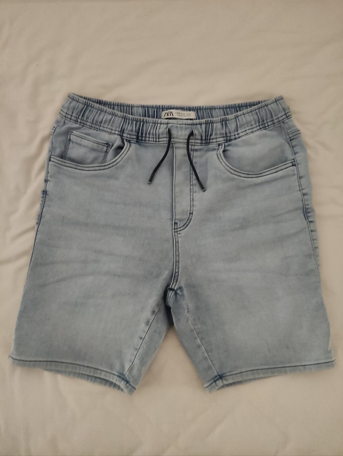 Light blue denim shorts with elasticated waistband and black drawstring, on a white surface. Label visible on waistband.