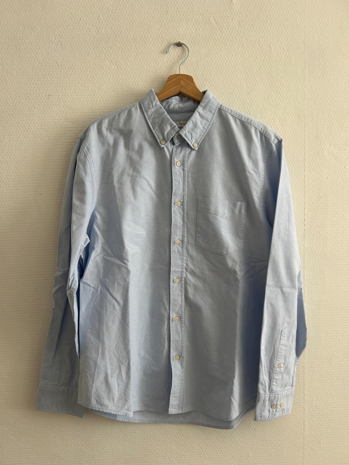 Light blue long-sleeved shirt hanging on a wooden hanger against a beige wall.