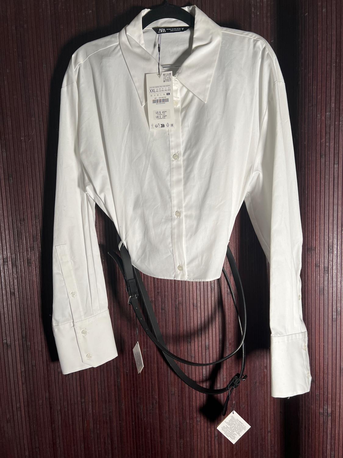 White long-sleeved shirt hanging on a hanger with a visible tag. Dark wood background. Black belt included.