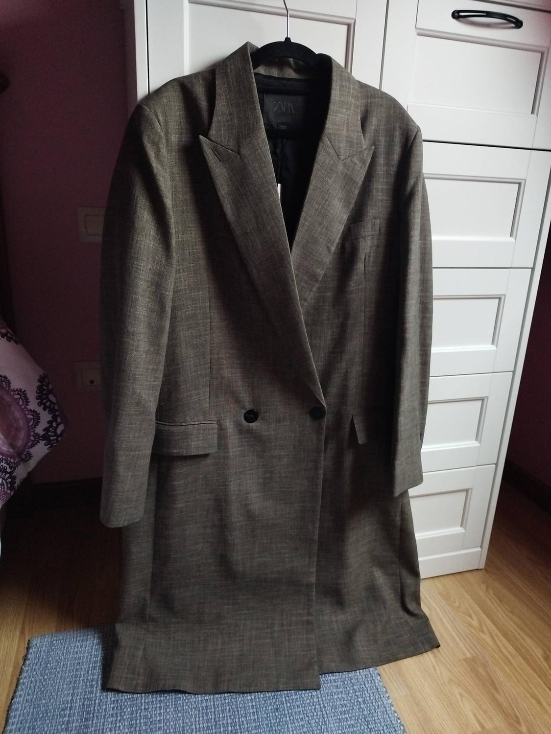 Long grey coat with black buttons hanging on a hanger, in front of a white wardrobe on a blue rug.