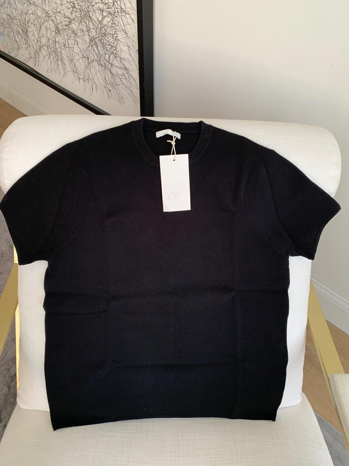 Black short-sleeved T-shirt with a hang tag, placed on a white armchair. Background with framed abstract art.