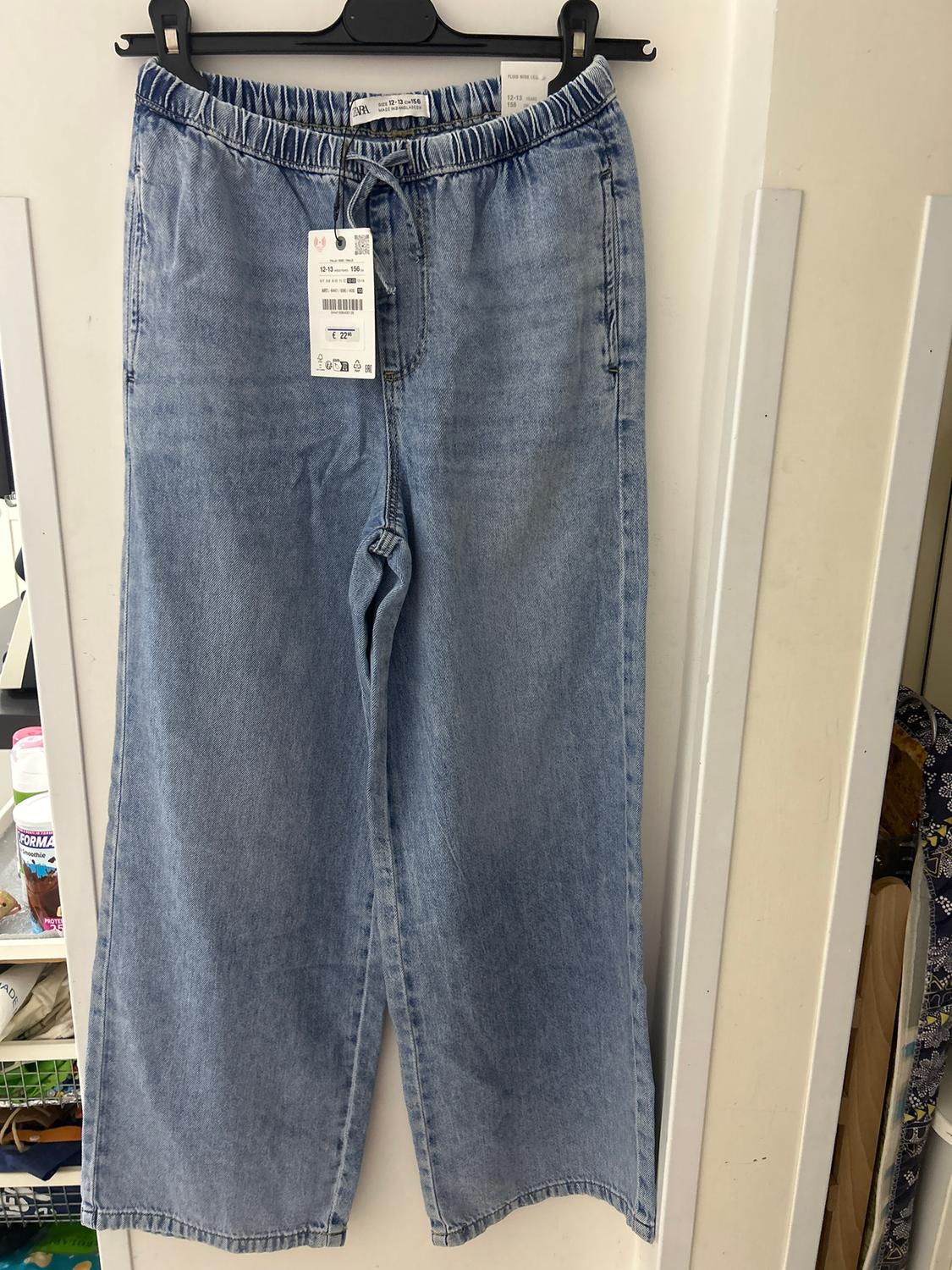 Light blue denim jeans hanging on a hanger, with price tag visible. White wall background and shelf with products.