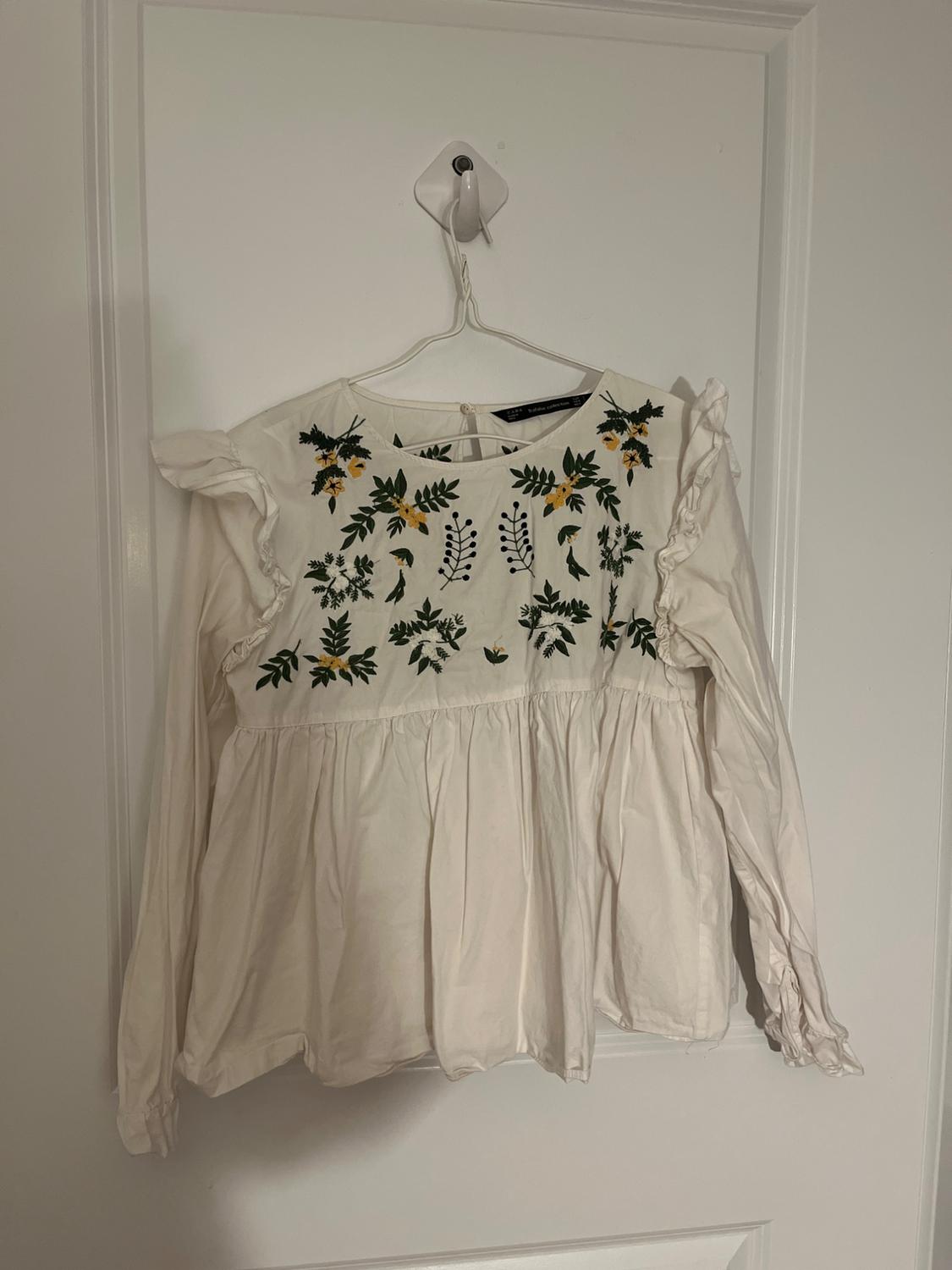 Front view of a white blouse with floral embroidery on the chest and long, ruffled sleeves.