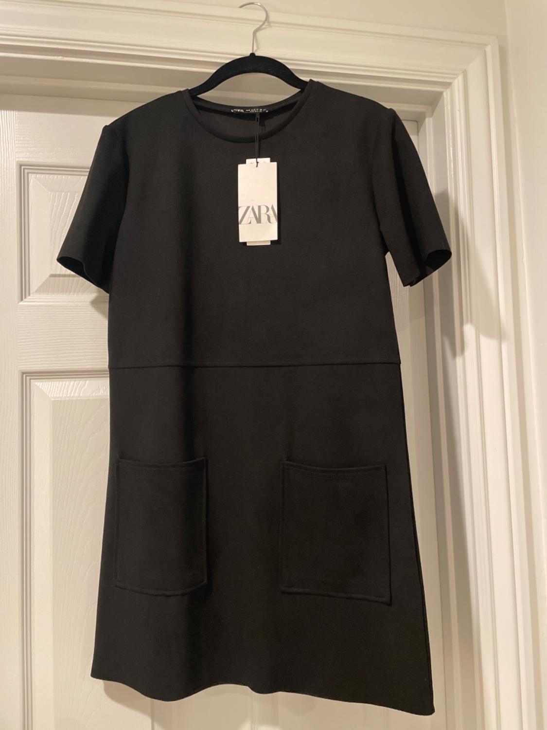 Black short-sleeved dress with two front pockets, hanging on a black hanger over a white door. Visible label with Zara logo.