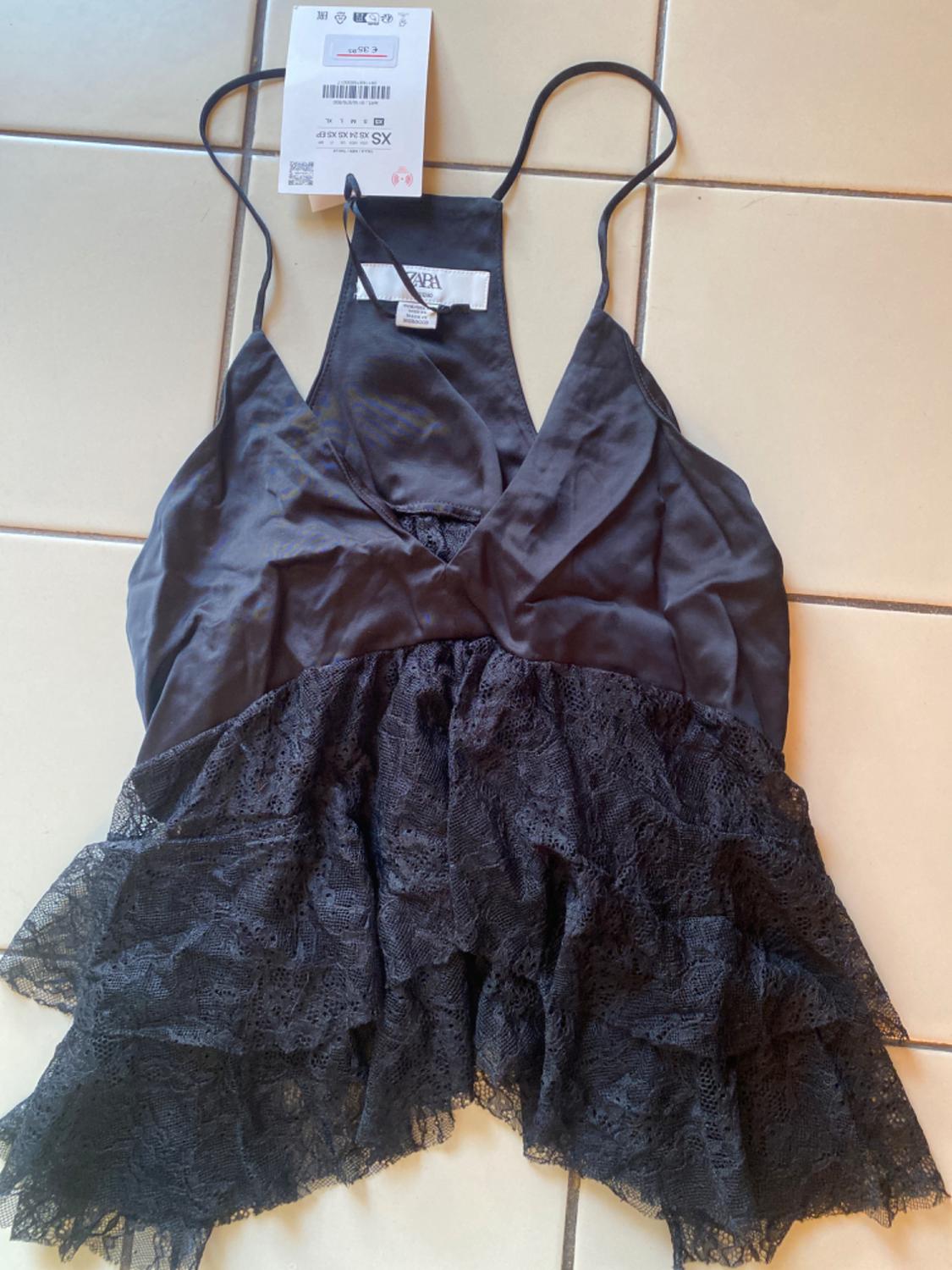 Black camisole top with V-neck. Smooth upper half, lower half with lace ruffles. Text: ZARA XS € 35.95. On tiles.
