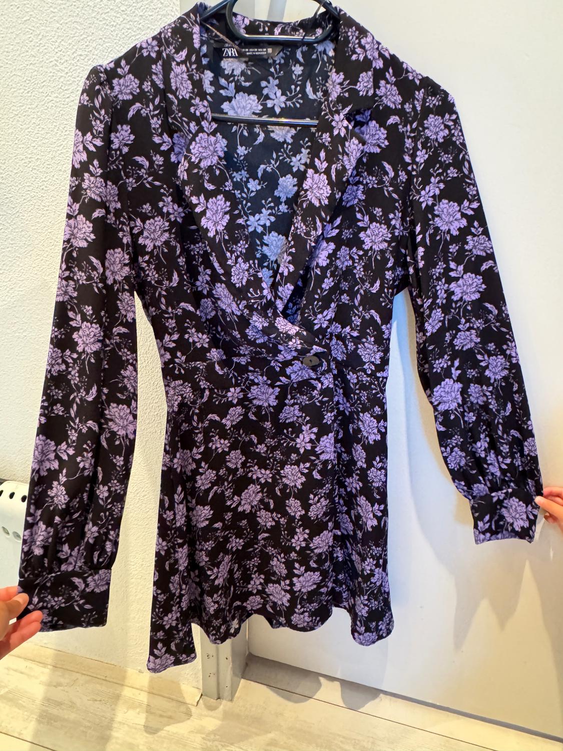 Short black dress with a purple floral print, long sleeves, hanging on a clothes hanger. White background and light wood floor.