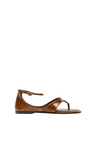 Brown patent leather sandal with ankle strap and flat black sole, on a white background.
