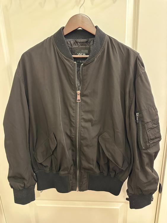 Black bomber jacket hanging on a wooden hanger. Front zipper detail and side pockets. "ZADA" label visible.