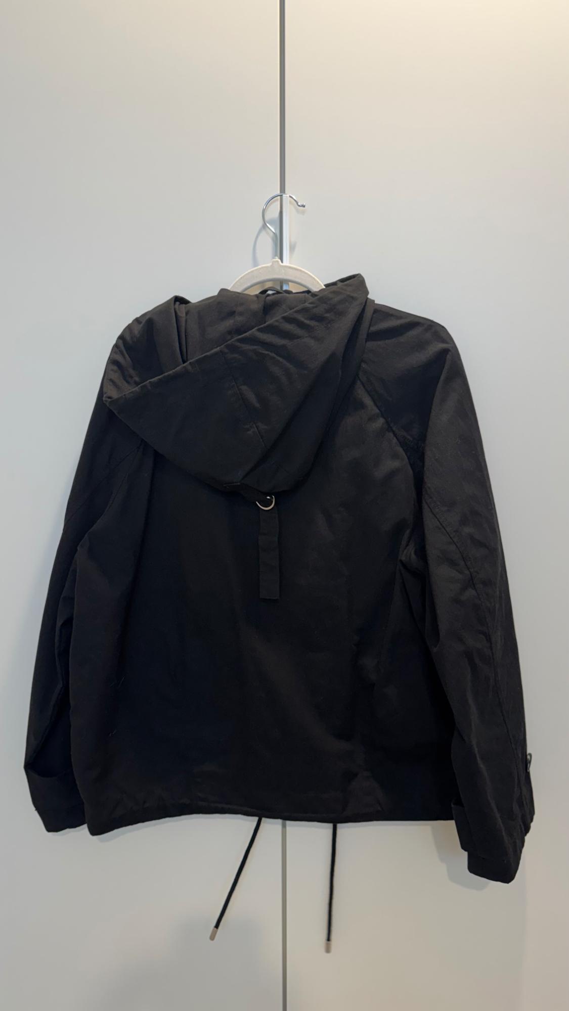 Black hooded jacket hanging on a white hanger against a light background. The jacket has drawstrings at the bottom.