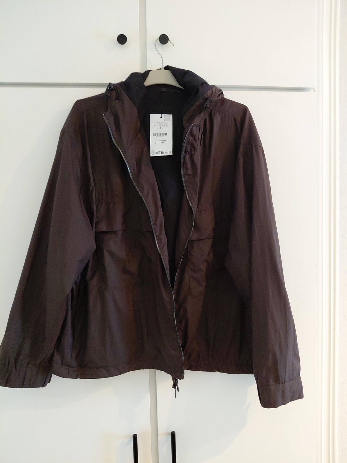 Brown jacket with a hood hanging on a hanger in a white wardrobe with black handles. It has an open zip and pockets.