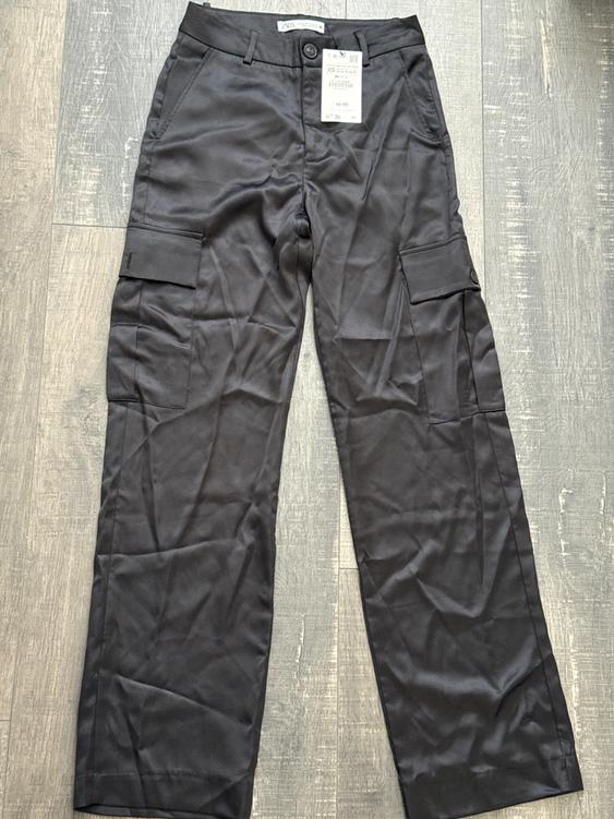 Black straight-cut pants with side pockets, visible hang tag, on gray wooden floor.
