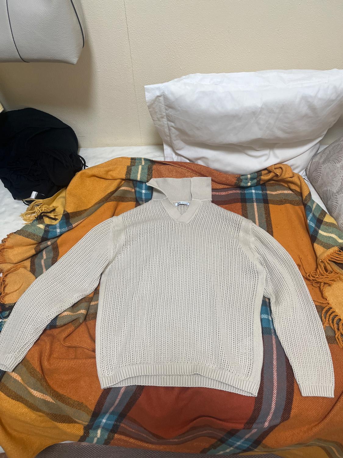 White knitted jumper on a tartan blanket in orange and blue tones, with white pillows and a grey handbag in the background.