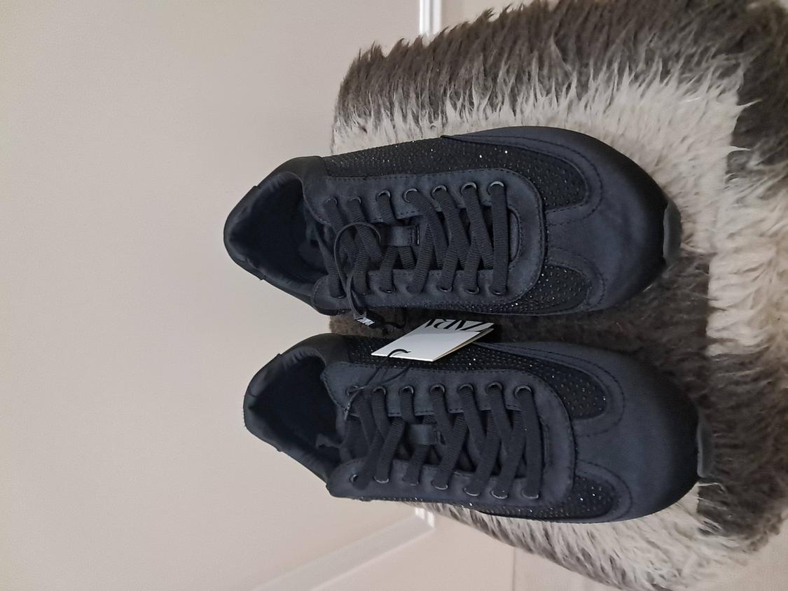 Black trainers with laces on a fluffy grey rug. Visible label with "ANDY" text. White wall background.