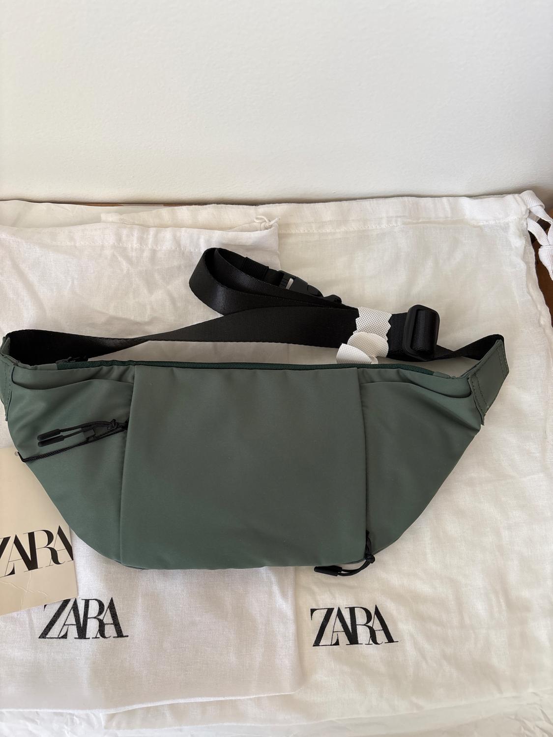 Dark green fanny pack with black strap on white fabric with the "ZARA" logo repeated. White background.