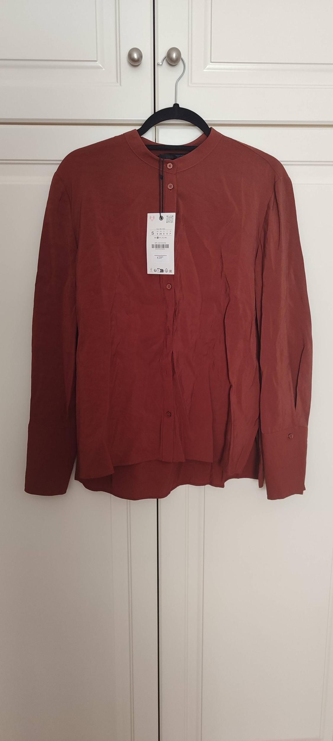 Brown long-sleeved shirt hanging on a black hanger in front of a white wardrobe. Visible tag with product details.