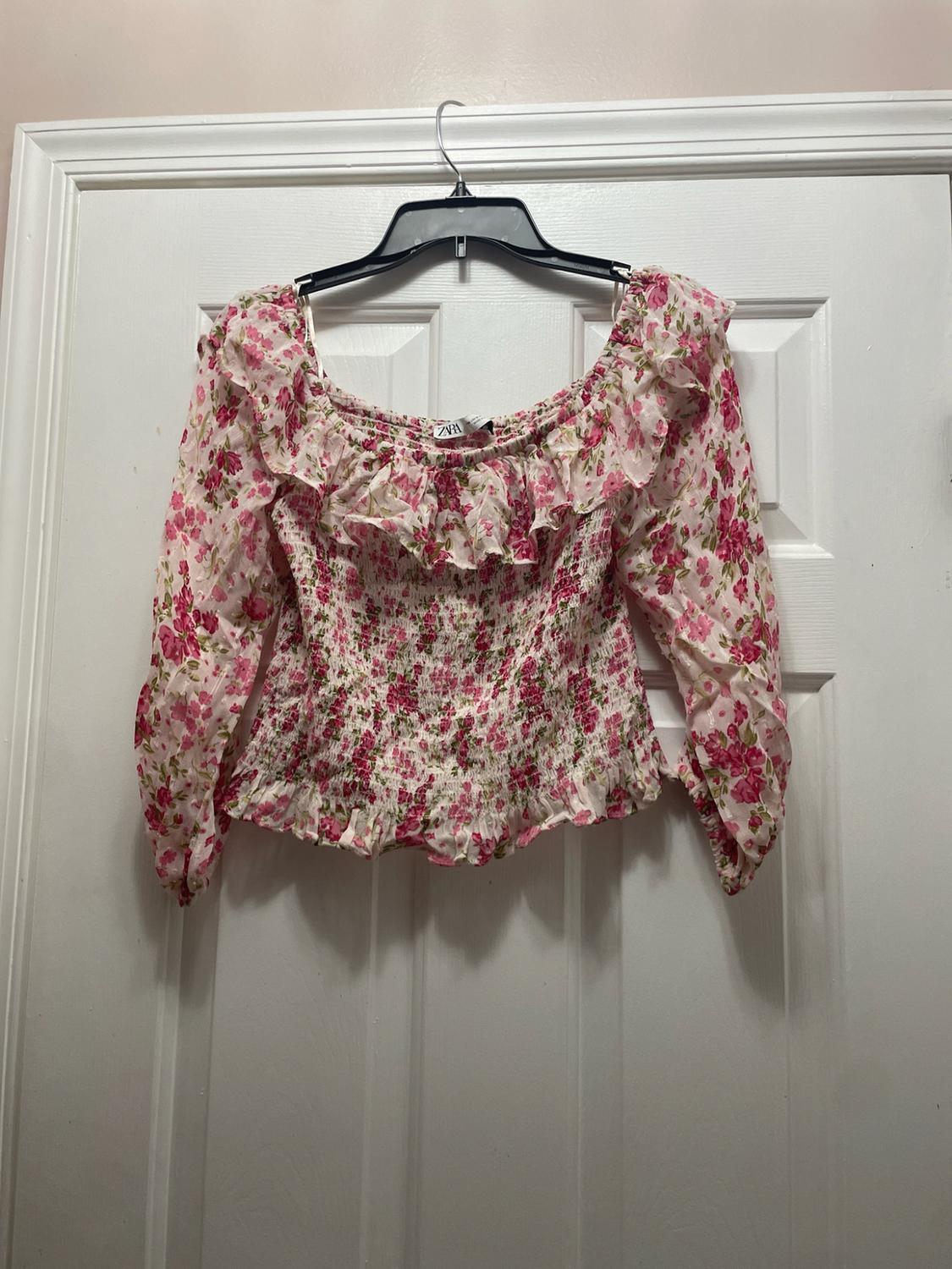 Long-sleeve blouse with a pink floral print, ruffled neckline, and shirred bodice. ZARA label. Hanging on a white door.