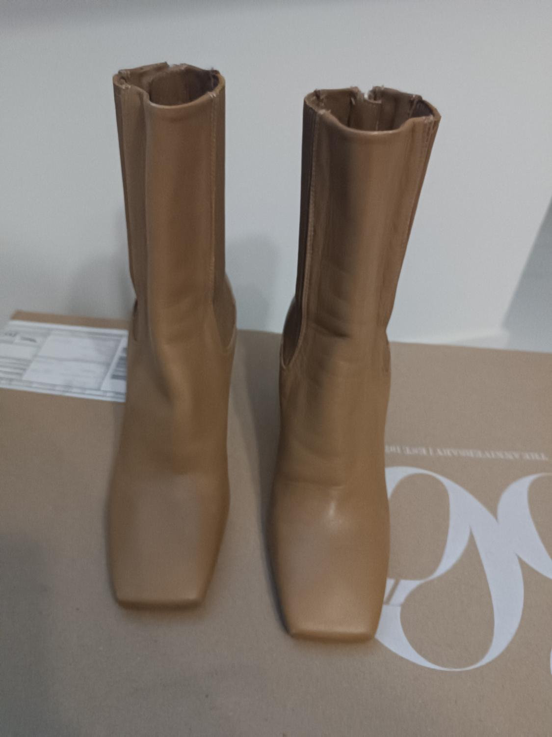 Beige knee-high boots with a square toe on a brown box with partially visible white lettering.