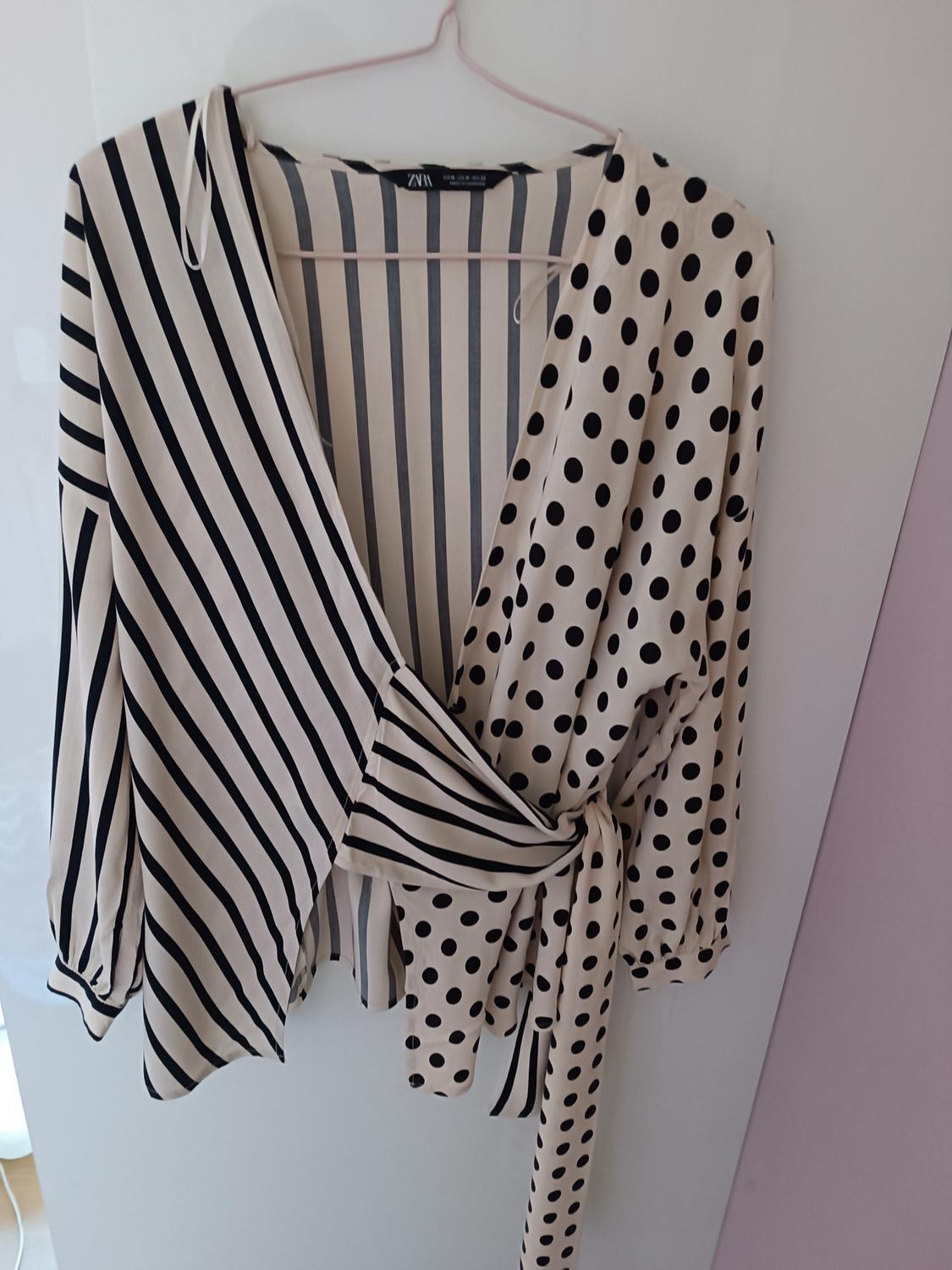 Blouse hanging on a hanger with a black and white striped and polka dot design, with a tie at the waist. White and lilac background.