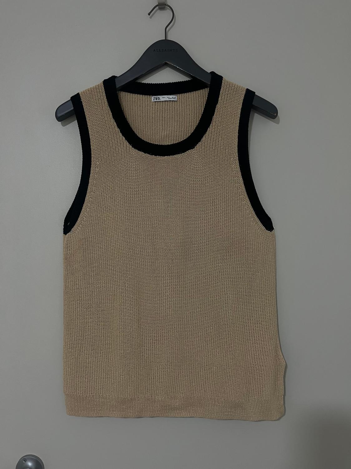 Beige sleeveless waistcoat with black trim, hanging on a black AllSaints branded hanger against a grey background.