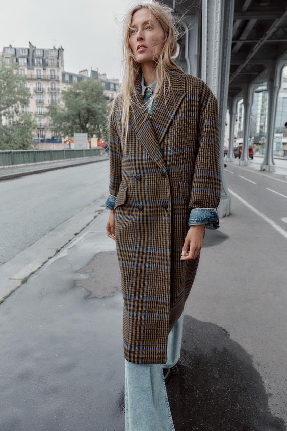 Souprava dlouhého hnědo-modrého kostkovaného dvouřadého kabátu s kapsami, přes světlé džíny.