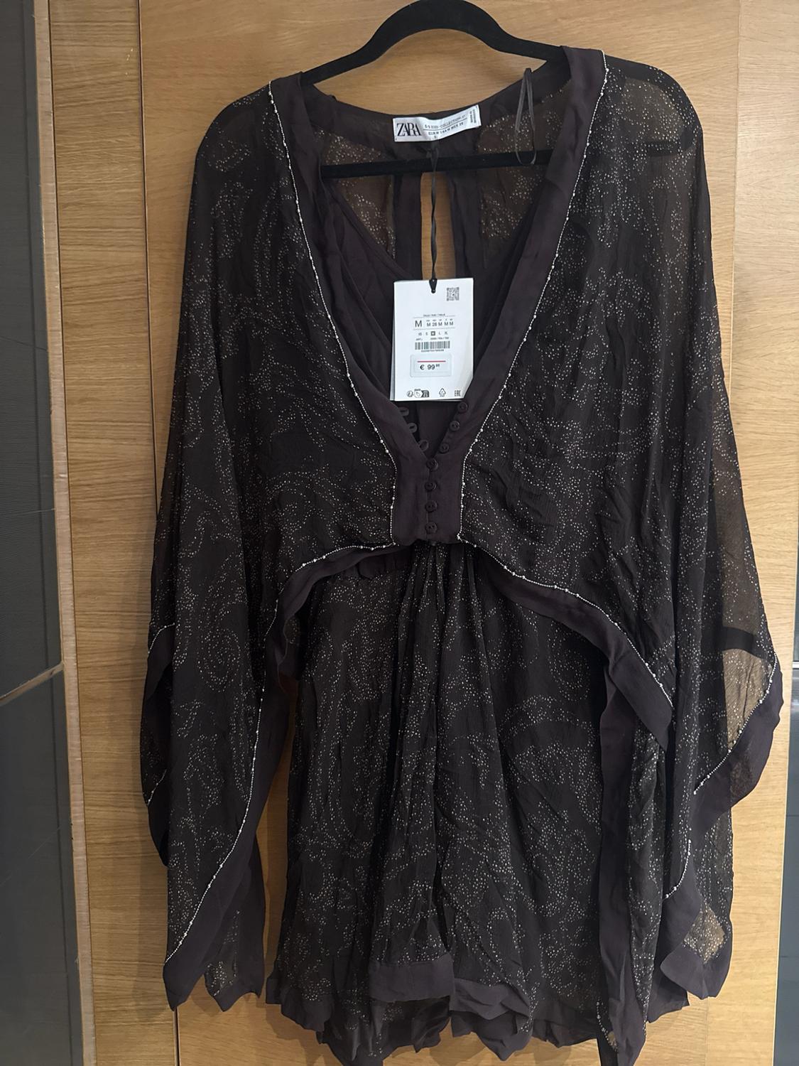 Semi-sheer dark garment with shiny details and wide sleeves hanging on a wooden door. Visible text: ZARA, M, €99.