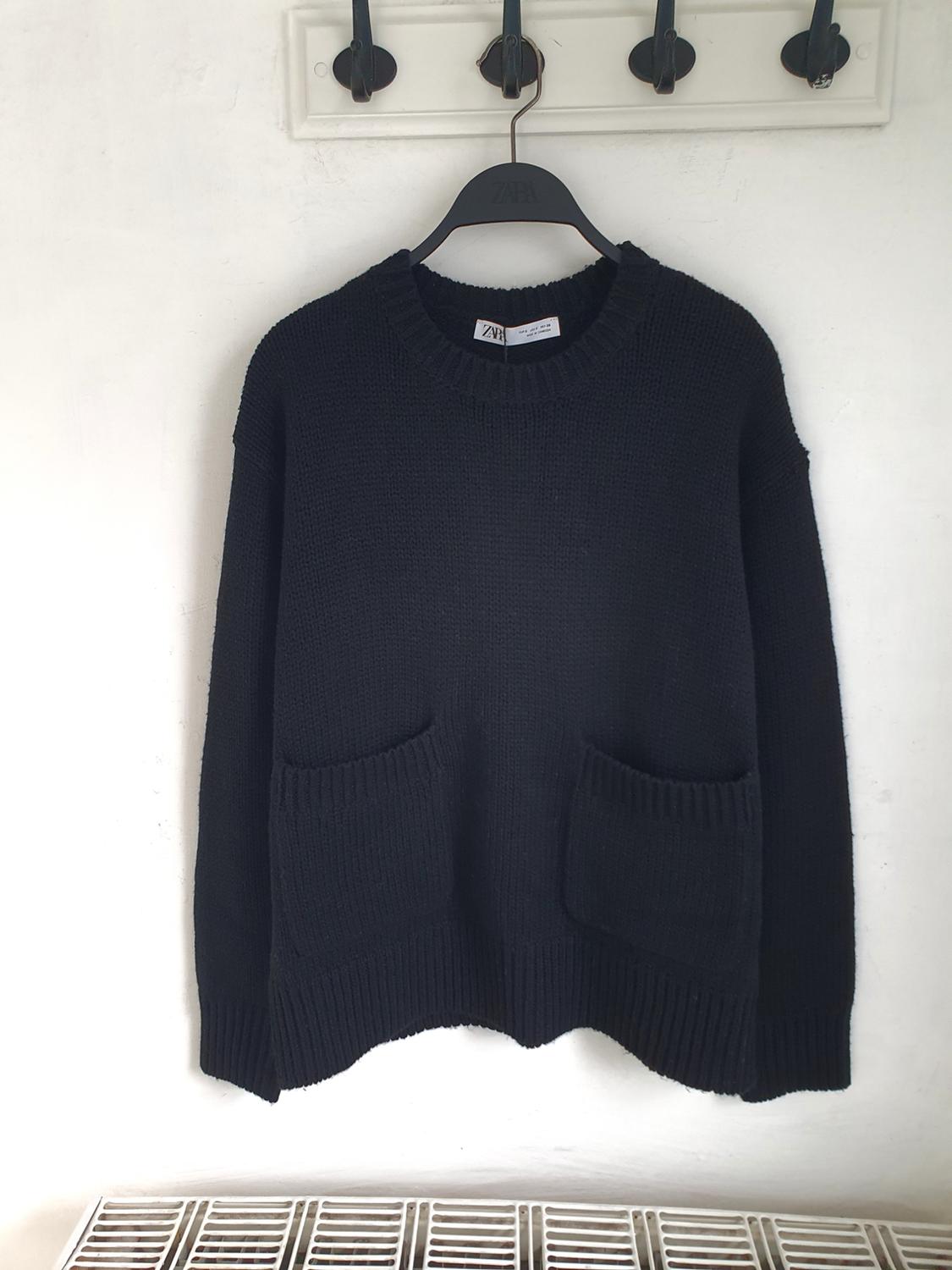 Chunky knit black jumper with a round neck and two front pockets, hanging on a black hanger against a white background.
