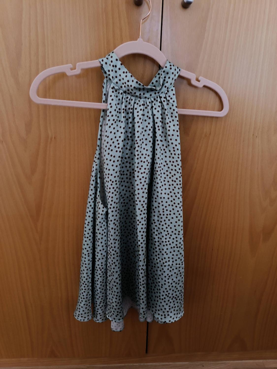 Ανοιχτό πράσινο top halter γιακά με σκούρες βούλες, κρεμασμένο σε μια ροζ κρεμάστρα μπροστά από ένα φόντο από ξύλινη ντουλάπα.