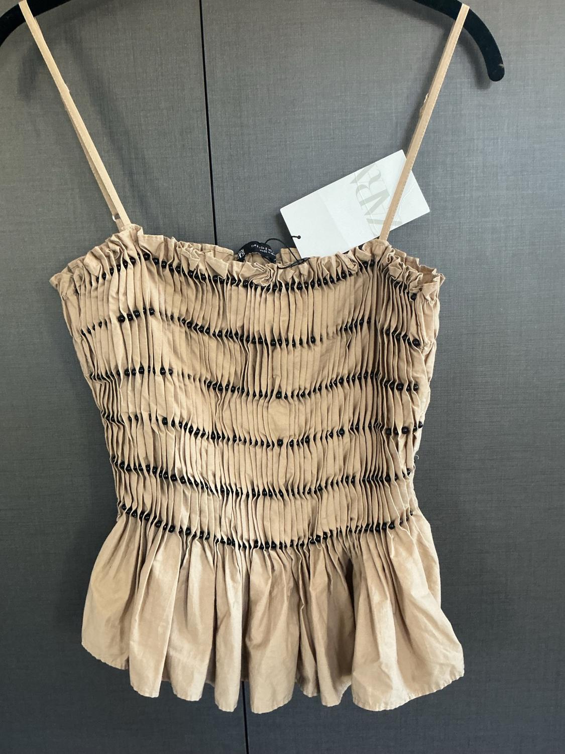 Beige camisole top on a hanger. Gathered bodice with black beadwork, flared hem, and a label with the text ZARA.