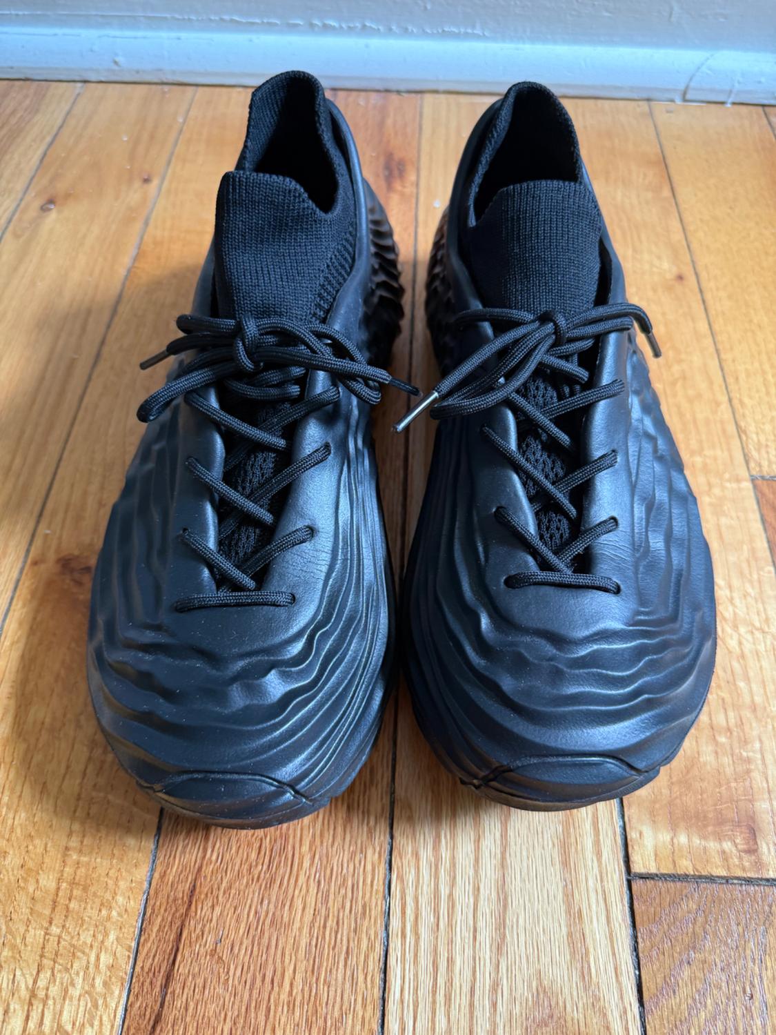 Black trainers with a modern design, featuring a wavy texture, laces, and a knitted collar, set on a light wood floor.