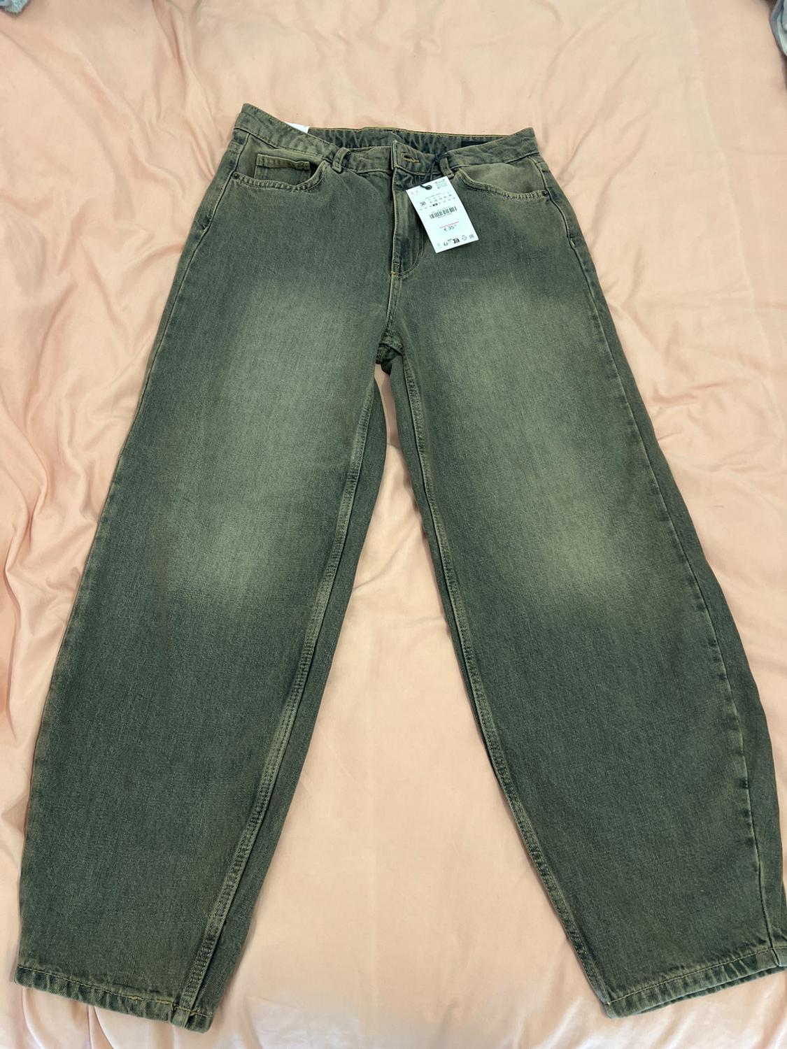Olive green wide-leg jeans on a bed with a pink sheet. White tag hanging from the waistband.