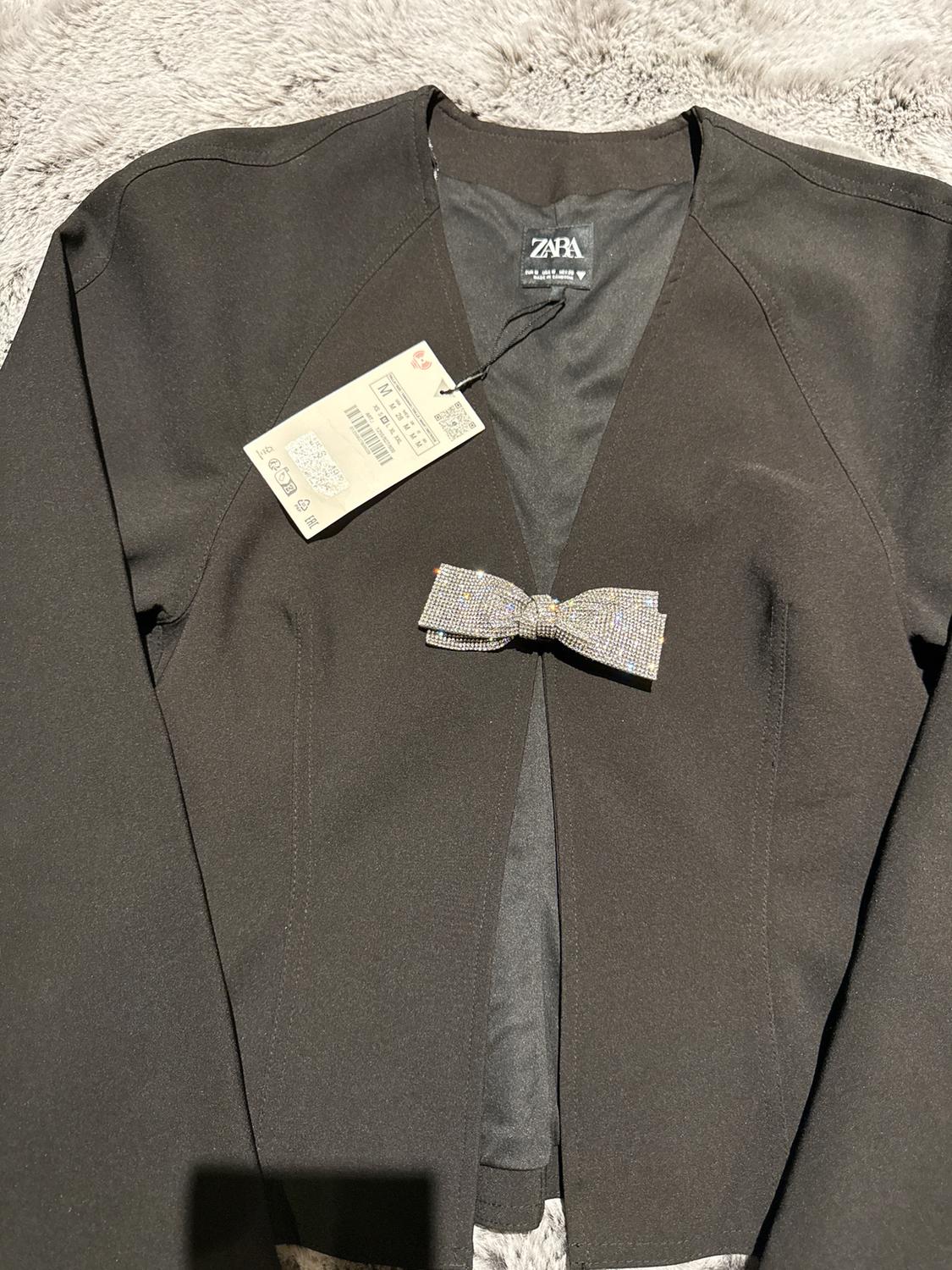 Black jacket with decorative bow on the front, Zara tag hanging. Fuzzy gray fabric background.