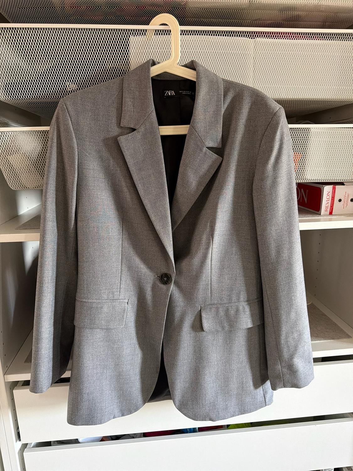 Grey blazer hanging on a hanger inside a white wardrobe. ZARA inner label. In the background, a box with the text REVLON.