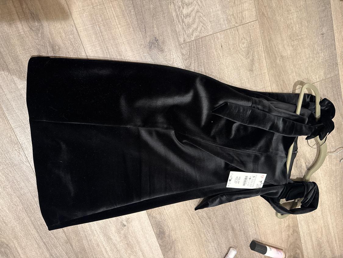 Black velvet dress on a wooden floor, hanging on a hanger. Visible tag with price and size details.