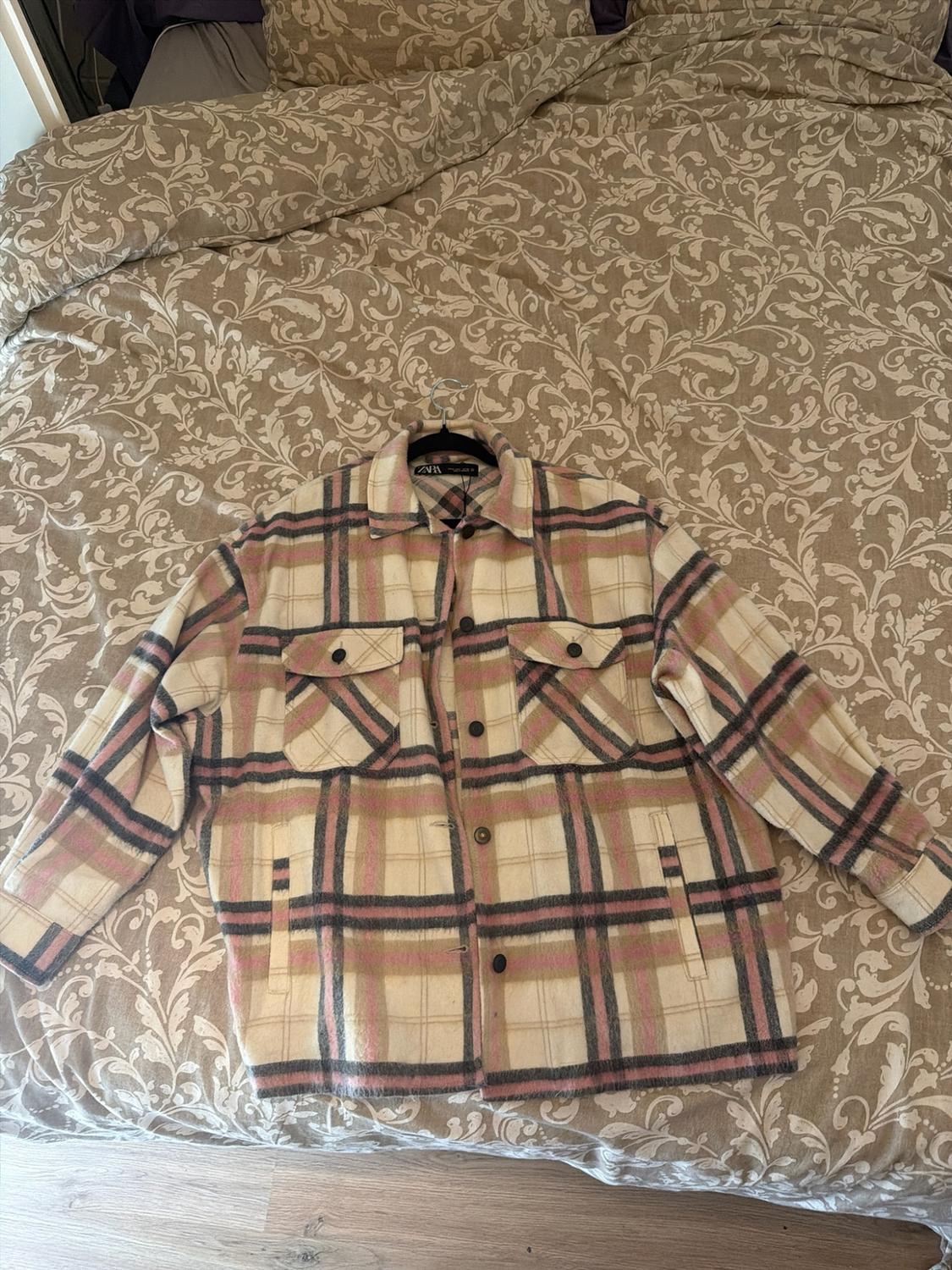 Beige, pink and grey checked jacket with pockets, on a hanger over a bed with a floral bedspread. ZARA label on the collar.