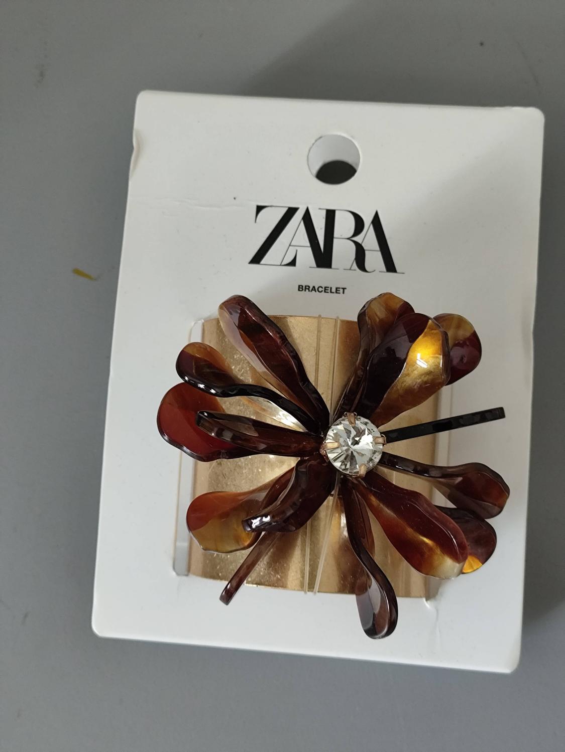 Gold-coloured bracelet with a tortoiseshell-effect flower and a central crystal, on a white tag with the text ZARA BRACELET on a grey background.