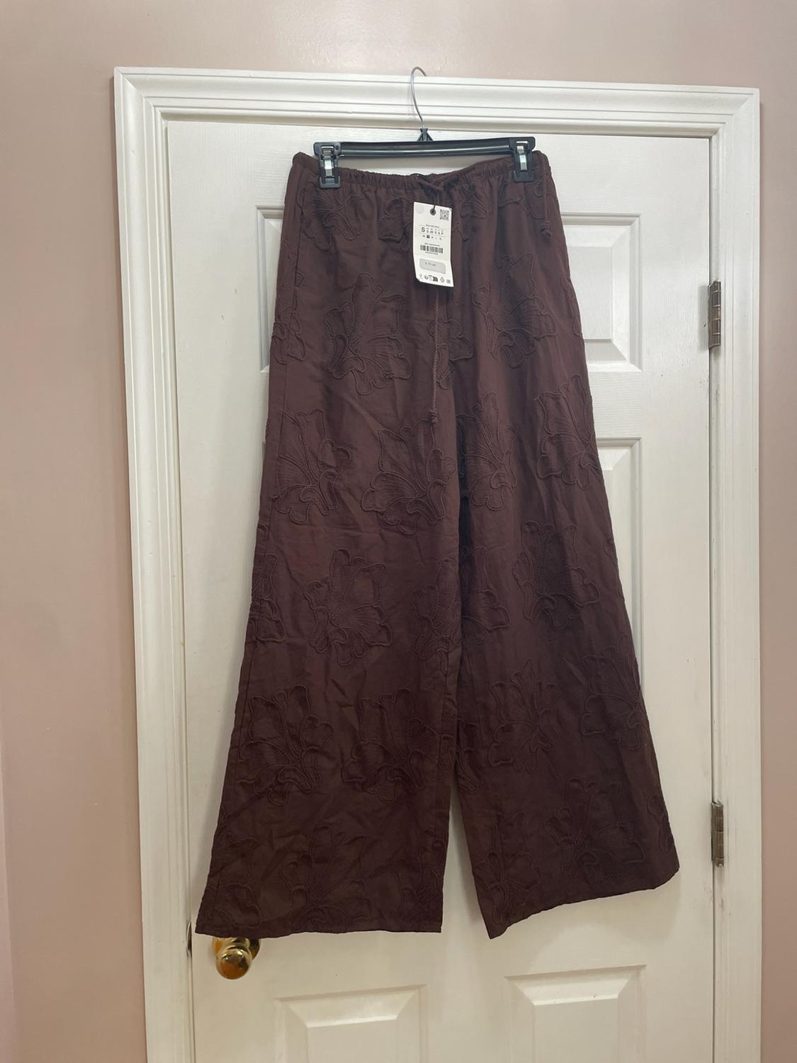 Wide brown pants with floral embroidery, hanging on a hanger over a white door. Elastic waistband with drawstring and visible label.