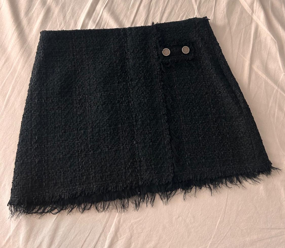 Textured black mini skirt with a frayed hem and two-button detail, resting on a crumpled white sheet.