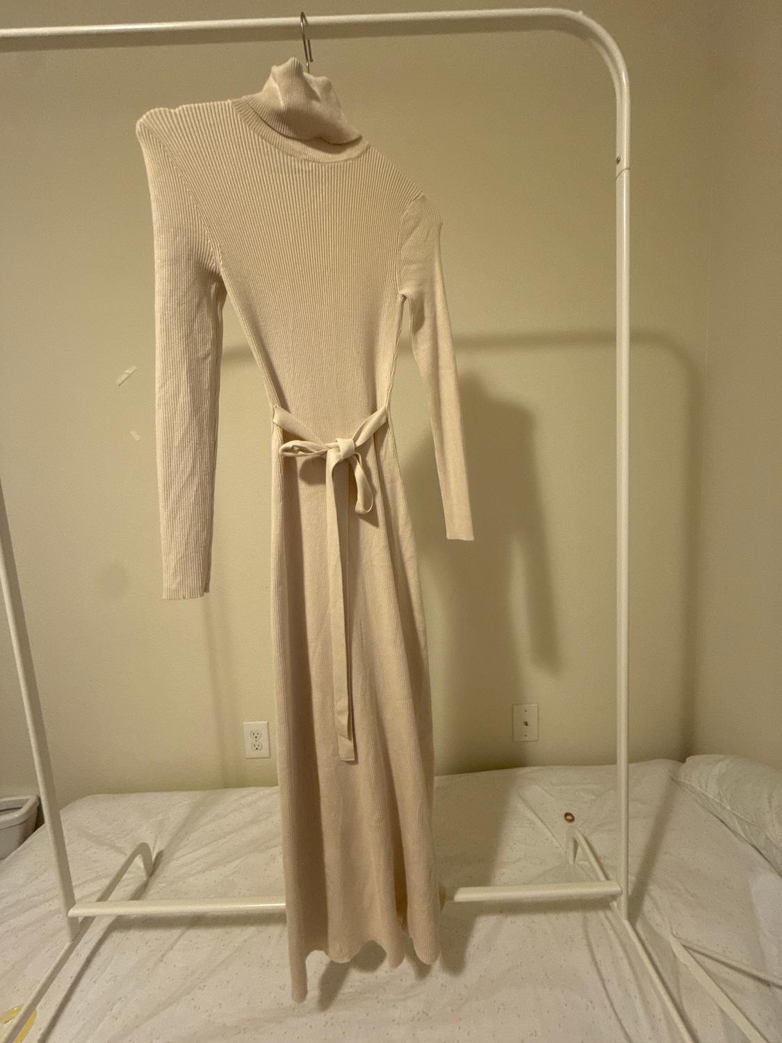 Long beige knitted dress with a high neck and belt, hanging on a white clothes rail over a bed.