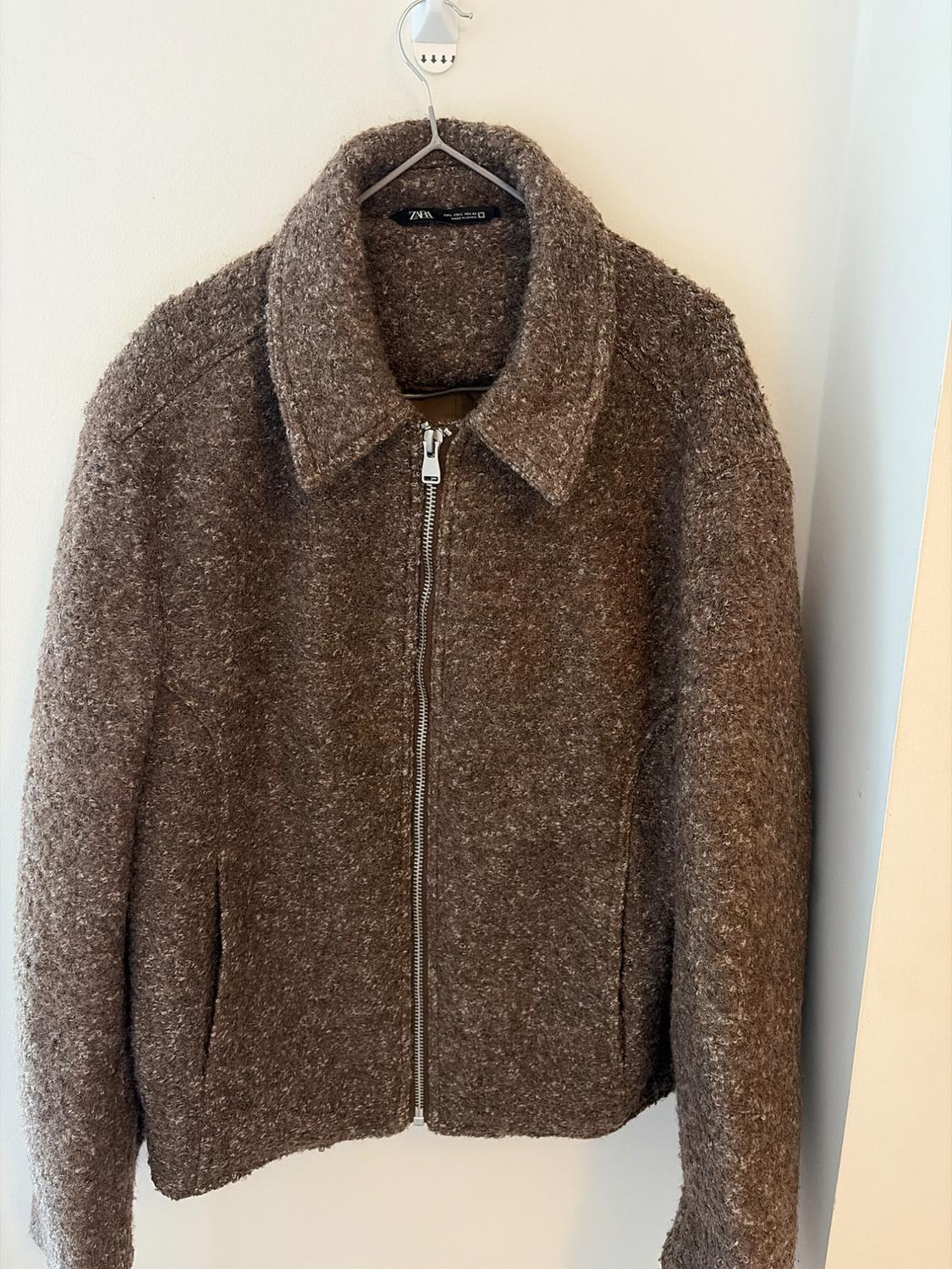 Textured brown jacket with collar and front zip, hanging on a hanger against a white wall. ZARA label.