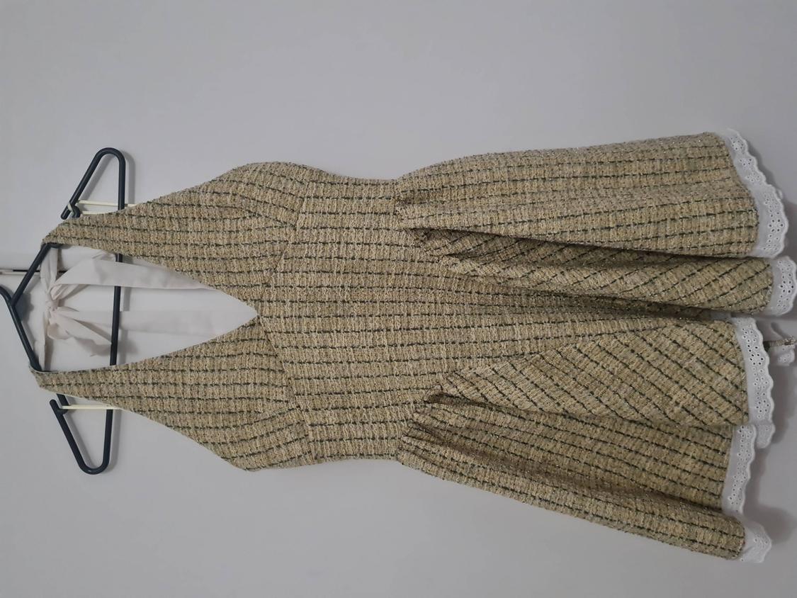 Short beige dress with dark checks, halter neck with ties and white lace hem, hanging on a hanger against a light background.