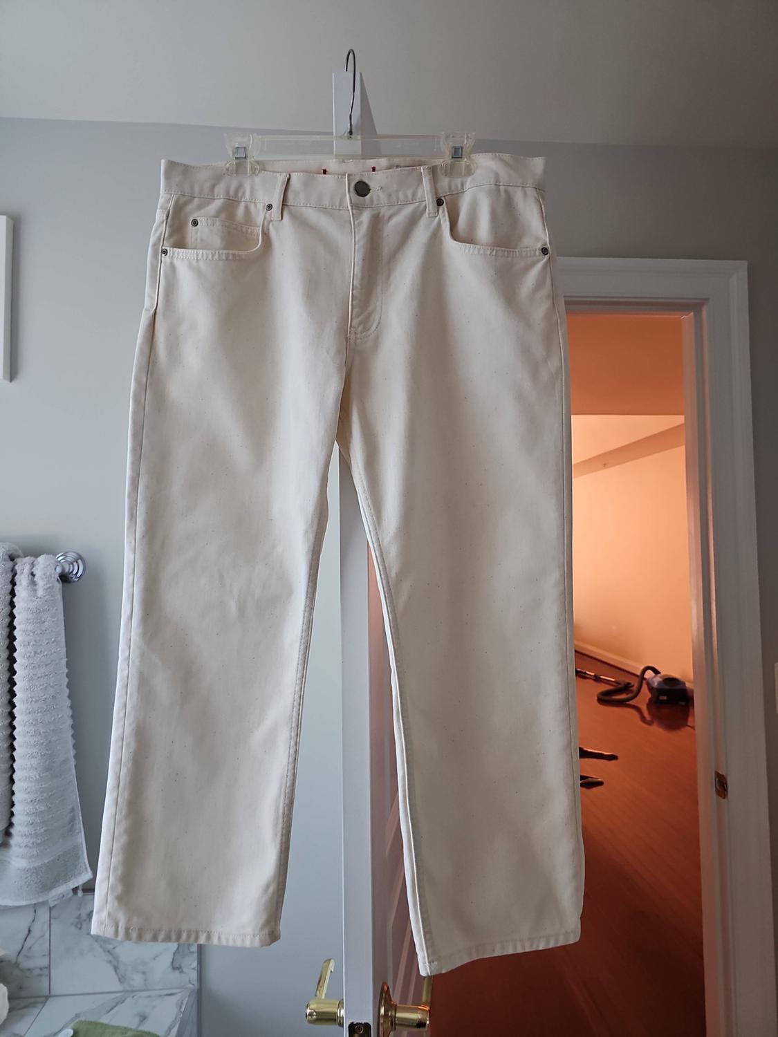Cream-colored denim jeans hanging from a plastic hanger in a bathroom, with a towel visible on the left.