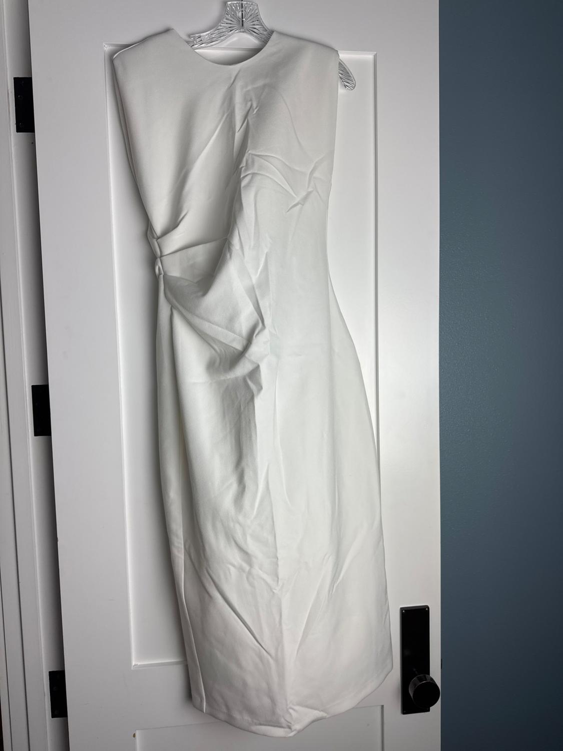 White sleeveless dress with drape detail, hanging on a hanger over a white door next to a blue wall.