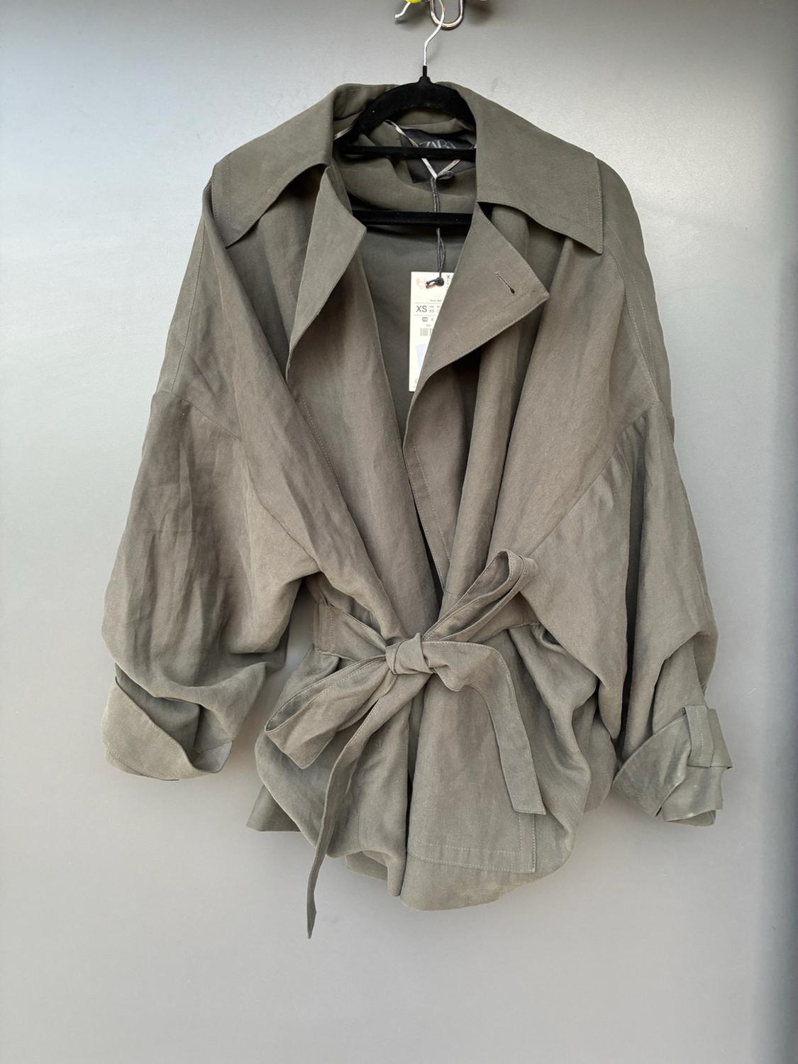 Olive-green jacket hanging on a black hanger against a grey background. It has wide sleeves, a tied belt and labels bearing the text ZARA and XS.