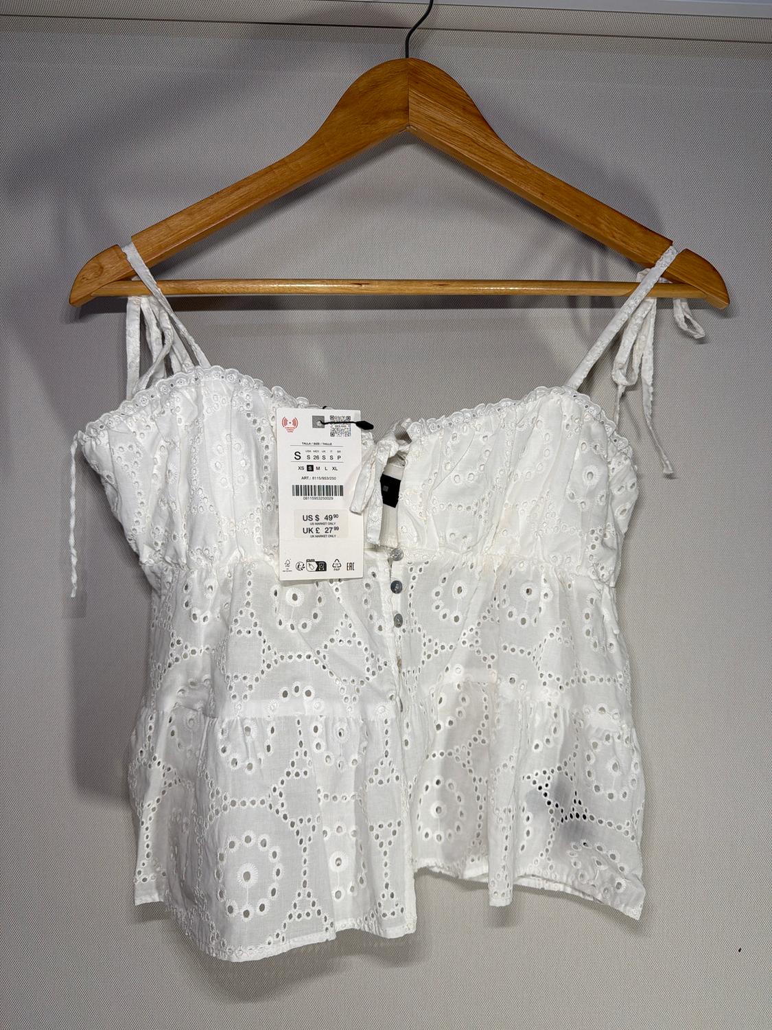 White strappy top with broderie anglaise, hanging on a wooden hanger. Visible tag with sizes and price.