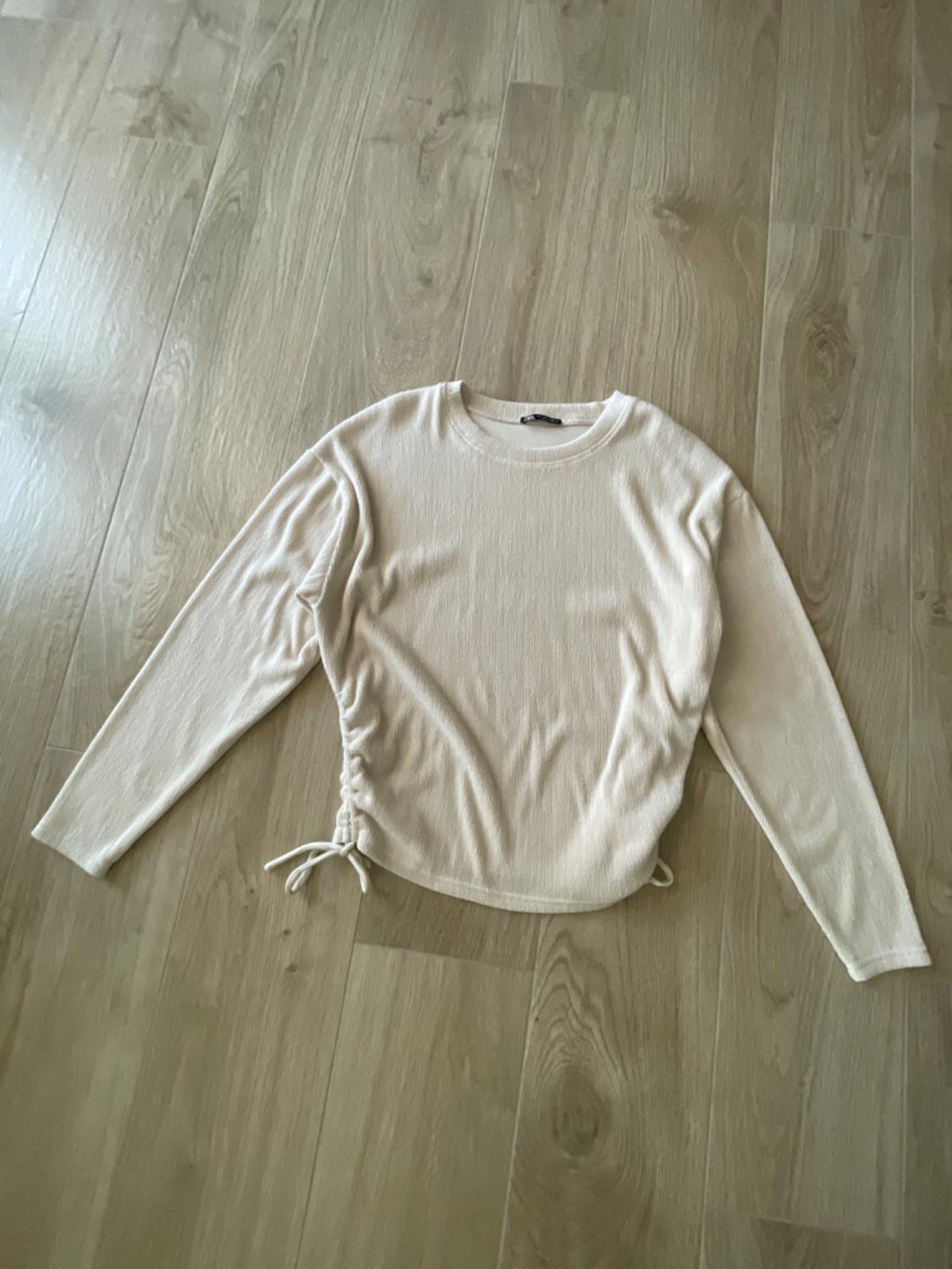 Long-sleeved beige jumper with side adjustment, placed on a light wooden floor.